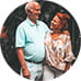 An elderly couple smiles and embraces while standing outside. The man wears a light blue shirt, and the woman wears a floral top. The scene conveys warmth.
