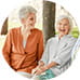 Two elderly women sitting outside on a bench, laughing and enjoying a conversation. They are surrounded by trees, conveying a warm and joyful atmosphere.